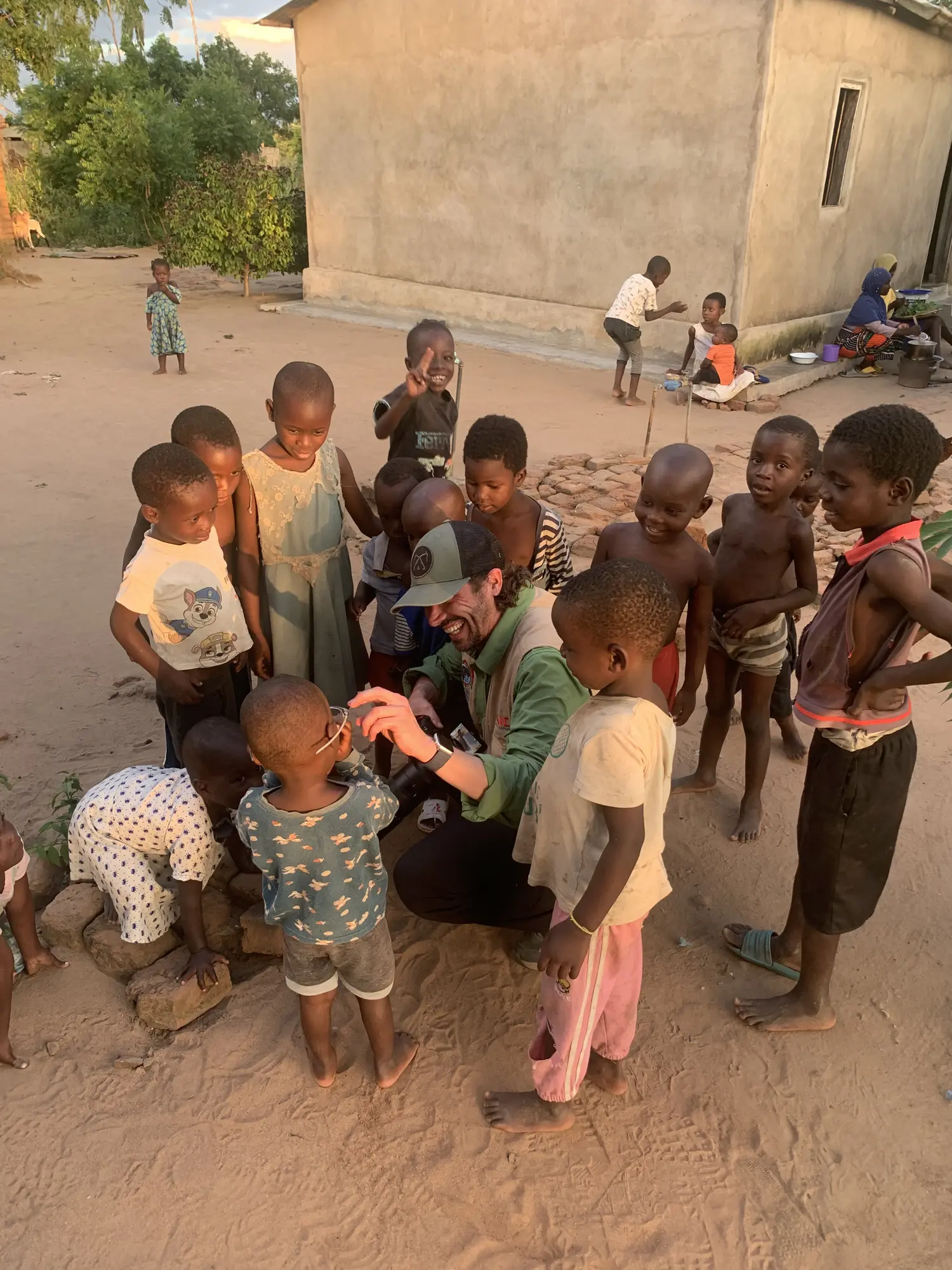 David crouches among children, smiling as a child tries on his glasses, with a camera in his lap