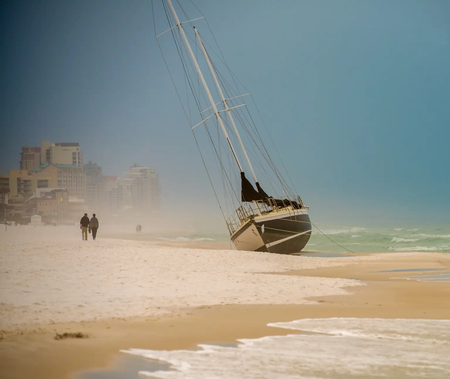 A beached sailboat leaning toward the surf as two people walk along a hazy Florida shoreline